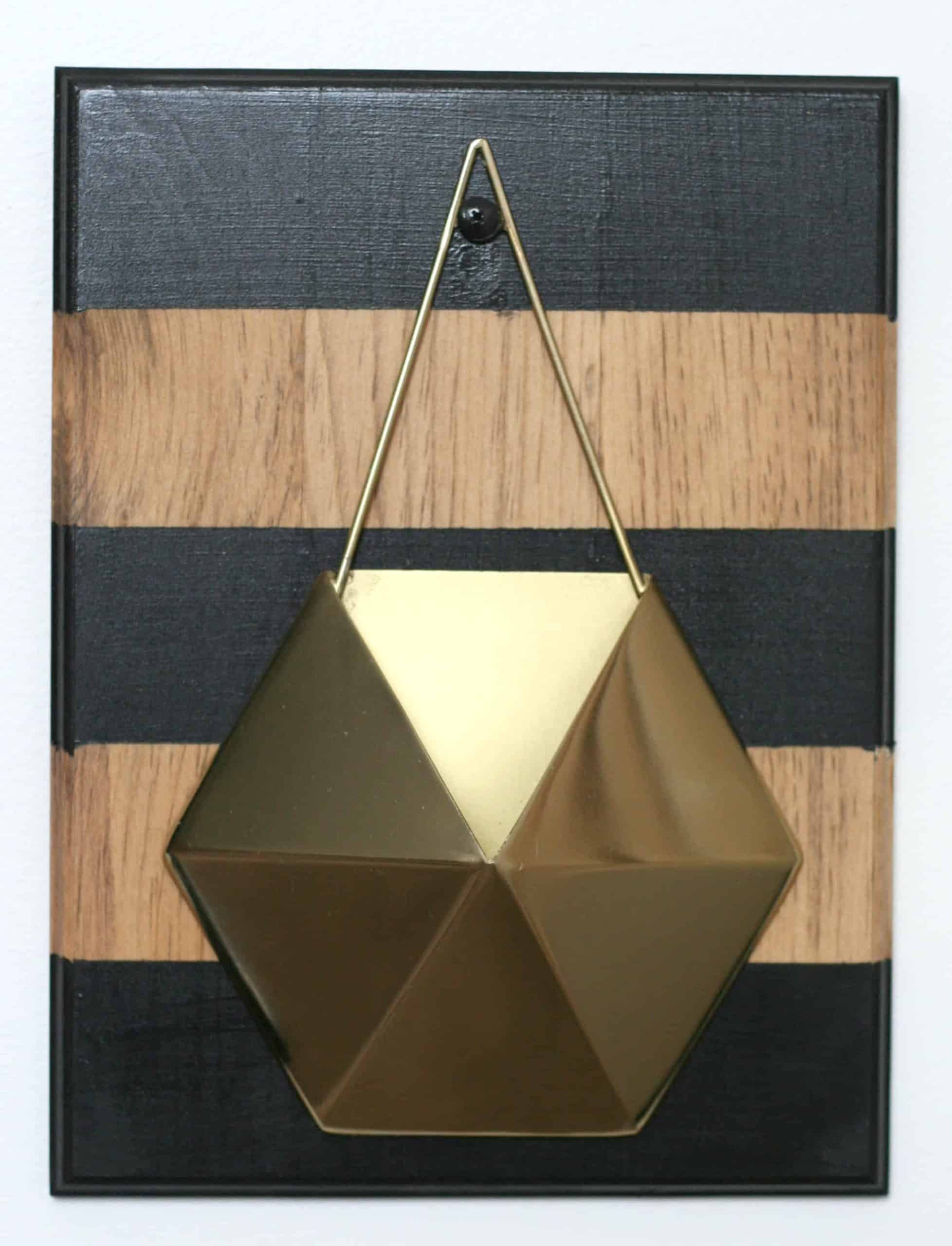 The completed geometric gold and wood wall planter. A hexagon-shaped gold planter hangs on a black and wood-striped pine board, hanging on a plain white wall.