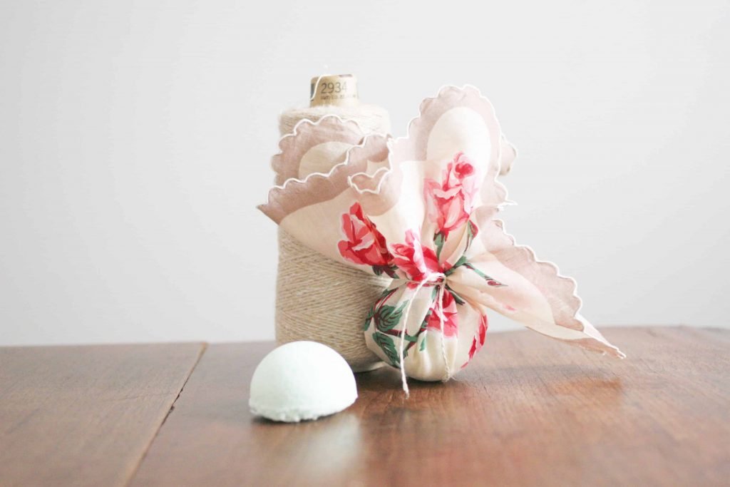 A set of homemade bath bombs sit on a wooden table top. Half of one bath bomb sits unwrapped on the table Another is wrapped in a tan linen, decorated with pink flowers, lace, and string.