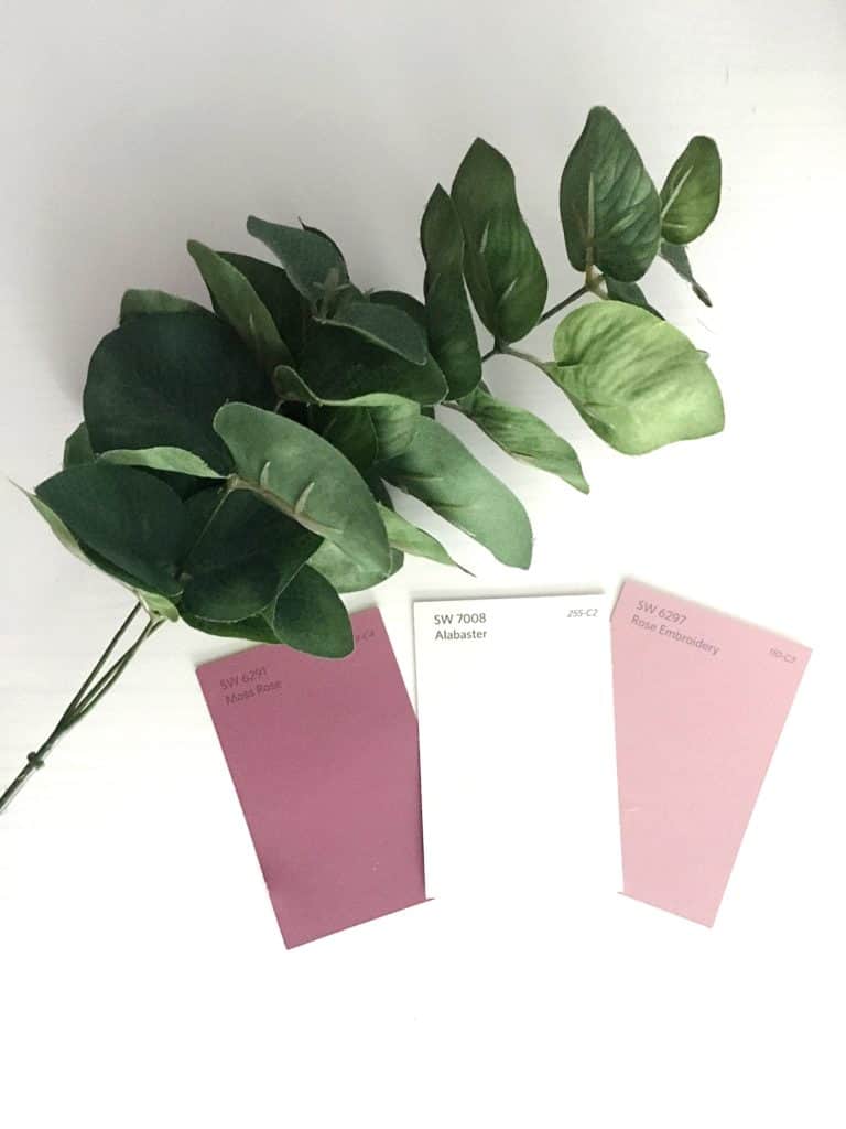 Three paint color cards lined up on a white surface - a mauvy rose color, alabaster white paint, and a light pink rose color. Stems of faux green leaves sit next to the paint cards.