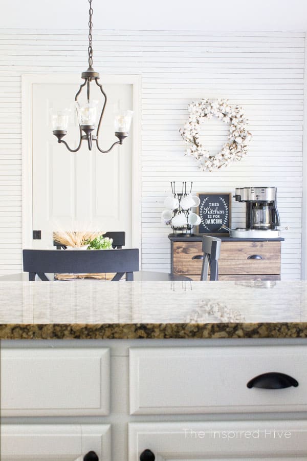 A view from a kitchen counter looks into a dinning room; the wall is covered in skinny shiplap, a dining table with chairs sits in front of a storage cabinet turned into a coffee bar with a mug holder and coffee maker.