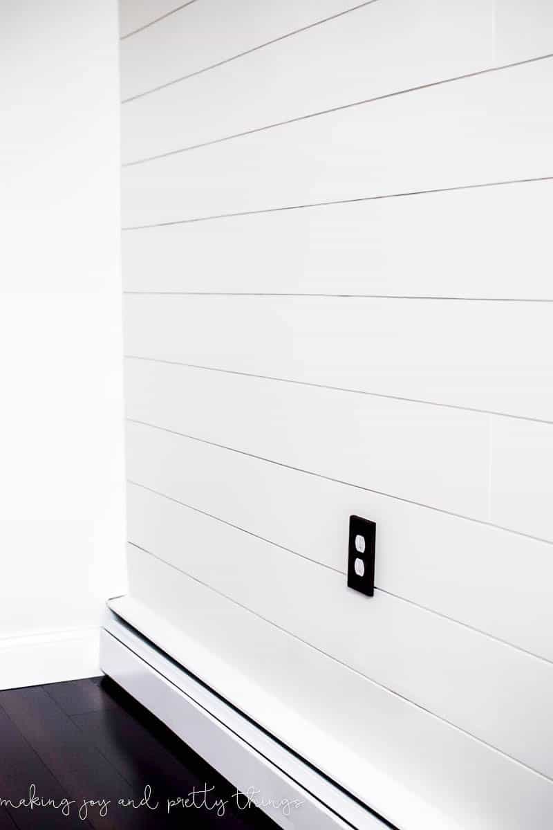 A white shiplap wall with a single electrical outlet. The outlet has a black plate cover.
