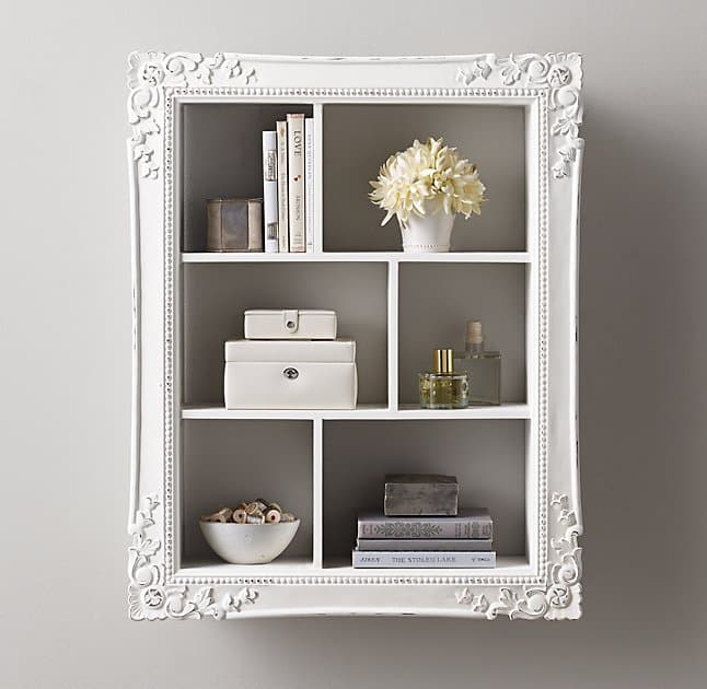 A white shadow box with ornate molding around the edges hangs on a white wall. The shadow box has6 different sized compartments, each holding different items; decorative books, faux plants, perfume bottles, and jewelry boxes.