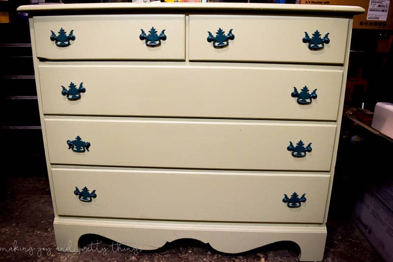 A 5-drawer dresser painted a plain cream color with ornate metal handles on each drawer.