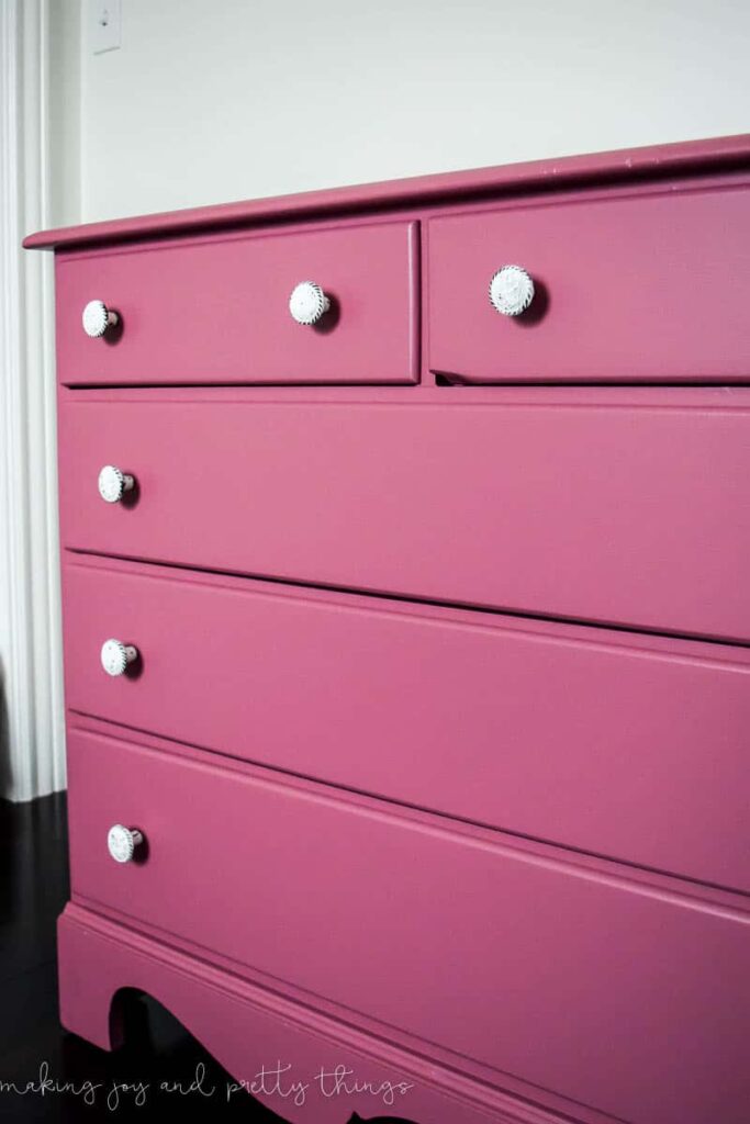 A side-angled view of the updated DIY pink dresser, painted a bold mauve pink. Each dresser drawer has a white knob handle.