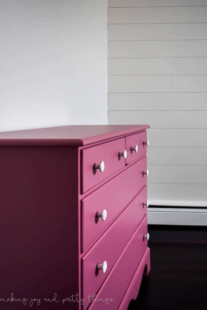 A side view of the DIY pink nursery dresser, freshly painted a dark mauve color with new knob drawer handles. The dresser is up against a plain white wall. In the background, there is a white shiplap wall.