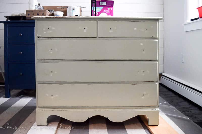 A 5-drawer dresser in the process of a DIY makeover. The drawer handles have been removed, there's spots of white spackle on the drawers, and the cream paint has been sanded down to show some of the natural wood tones underneath.