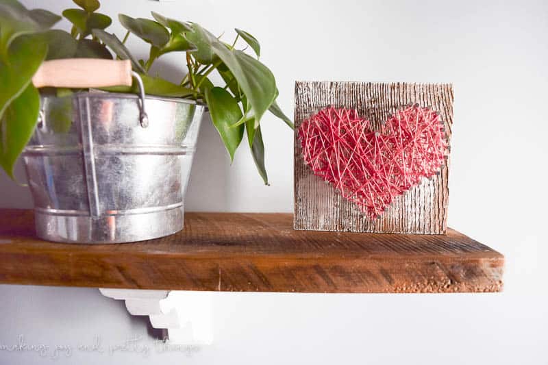 Small piece of barn wood with heart design using string art and nails sitting on floating shelf next to plant