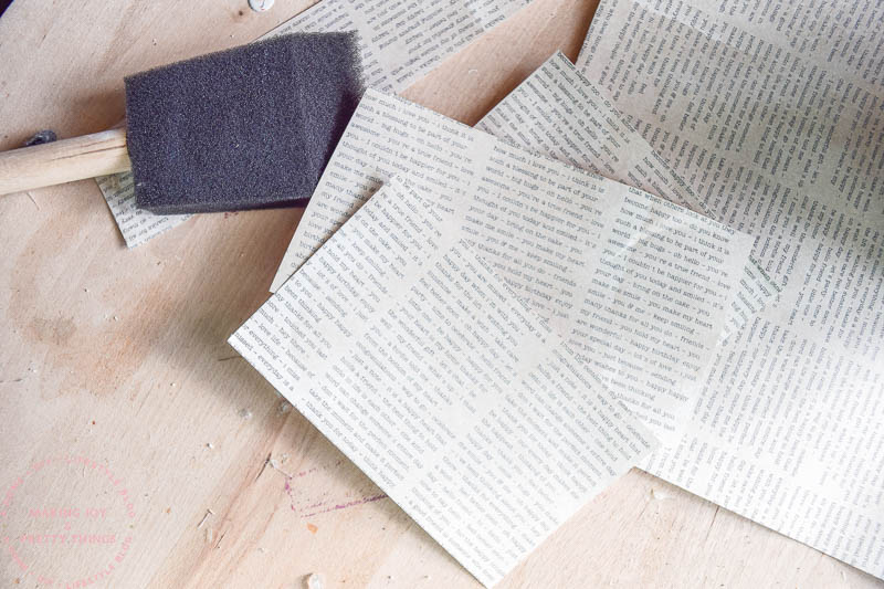 Pages of cardstock with lines of texts to look like book pages sit on a wood crafting table, next to a large crafting sponge.