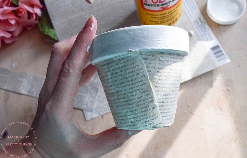 A hand holds a small terracotta pot that's painted white. Book page cardstock is decoupaged onto the sides of the planter.