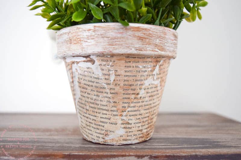 A closer look at the finished decoupage terracotta pots covered book pages. The rustic planter sits on a wood shelf and is filled with faux flowers.