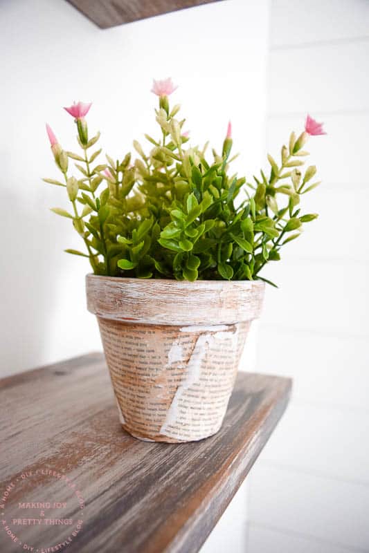 The completed decoupage terracotta pot sits on a wood shelf. The planter is filled with faux pink and green flowers.