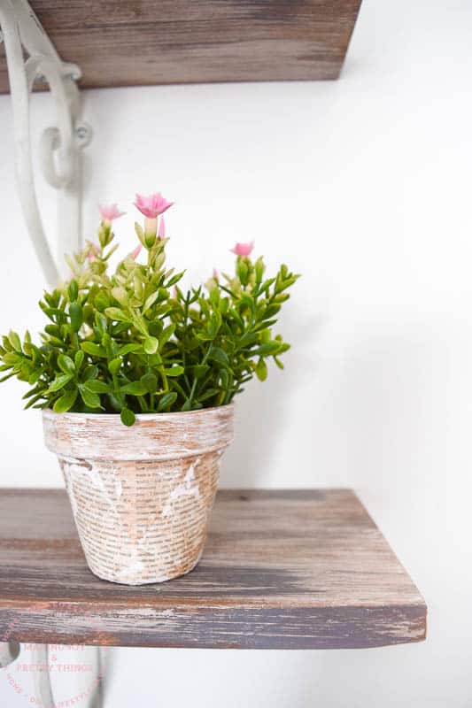 The completed decoupage terracotta pot made with old book pages sits on a rustic wood shelf. The planter is filled with faux pink flowers with vibrant green leaves.