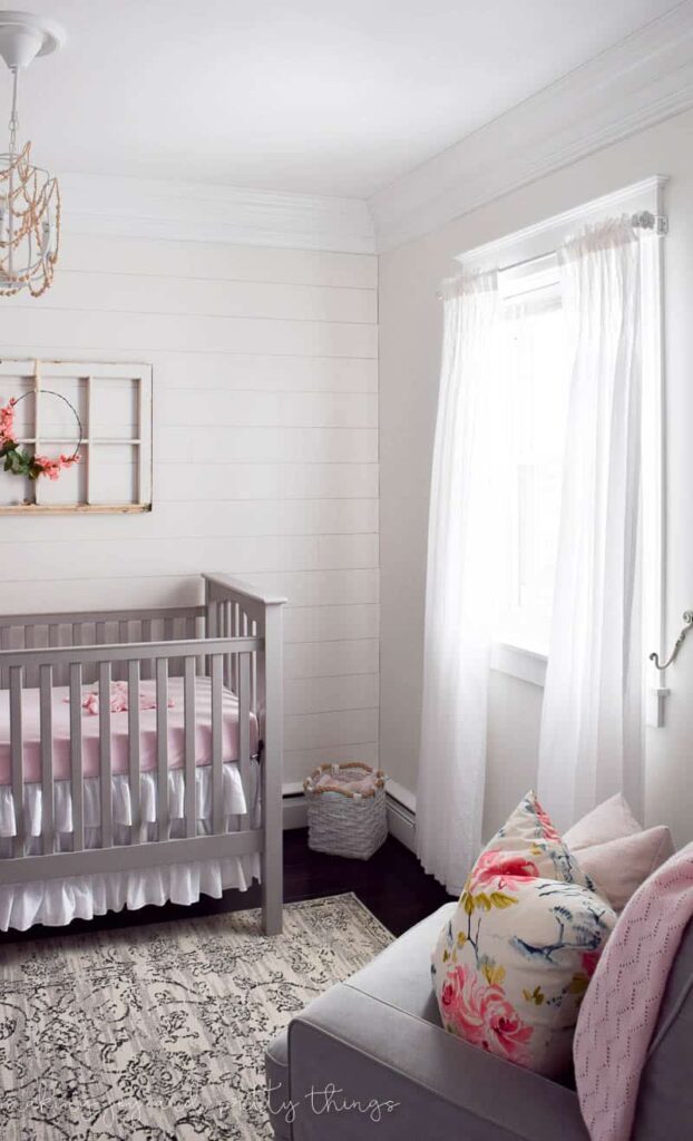 A little girl's nursery with white shiplap walls, a gray crib, and window-side seating area.