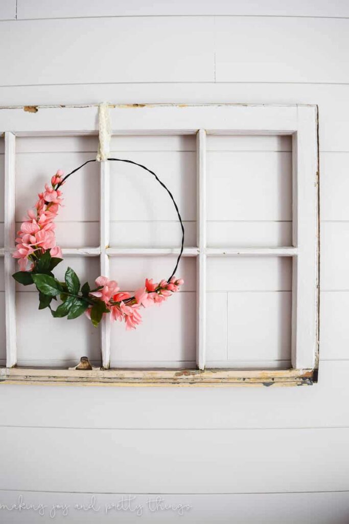 DIY floral wreath made with wire and faux flowers hung on a rustic window frame over a shiplap wall in a farmhouse nursery