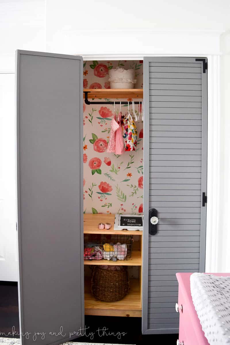 A peak into the closet in our baby girl's nursery, with shelves, wallpapered walls, and gray-painted shutter doors.