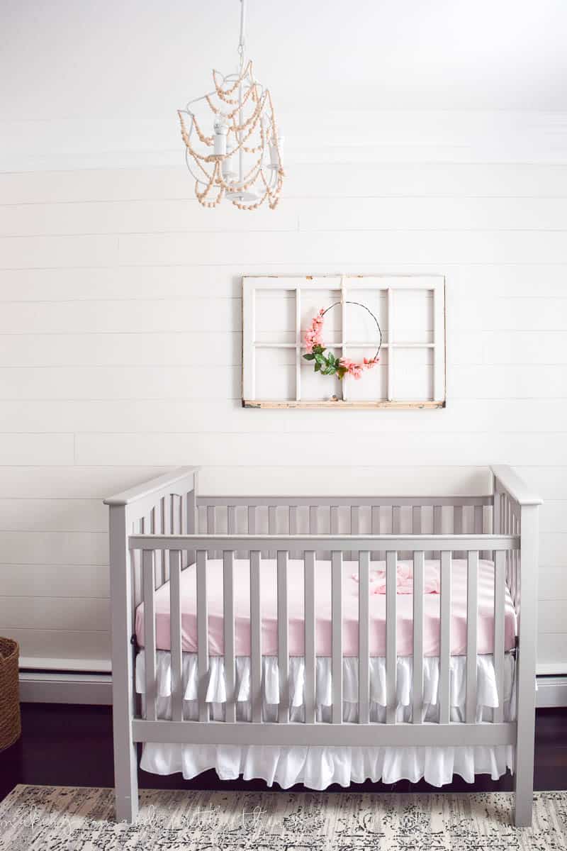 Antique window hung with a floral wreath done in rustic style over a crib on a shiplap wall with a wood bead lighting fixture