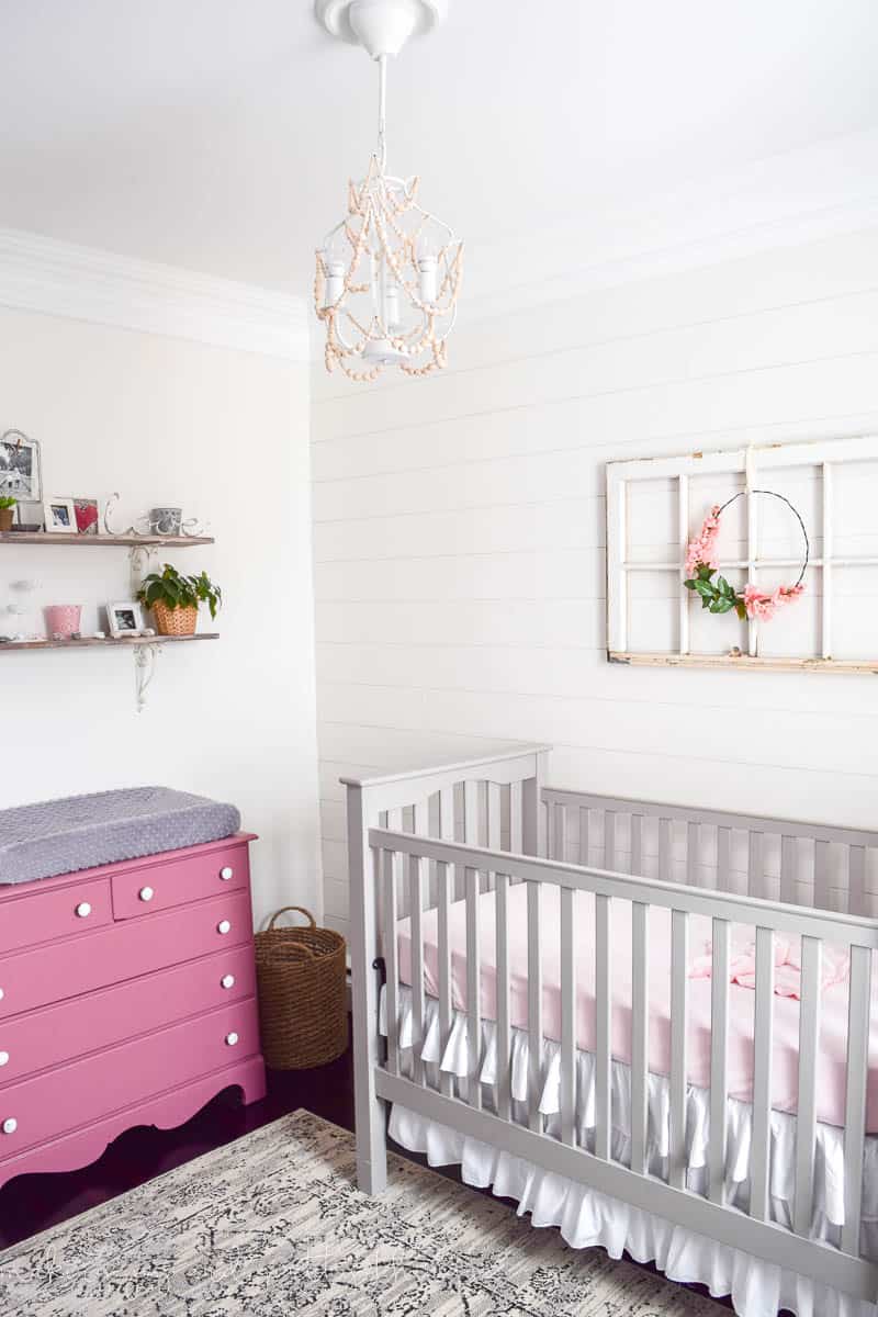 A crib, pink changing table and floating shelves, farmhouse style wall art, and diy wood bead chandelier in our baby girl's farmhouse nursery.