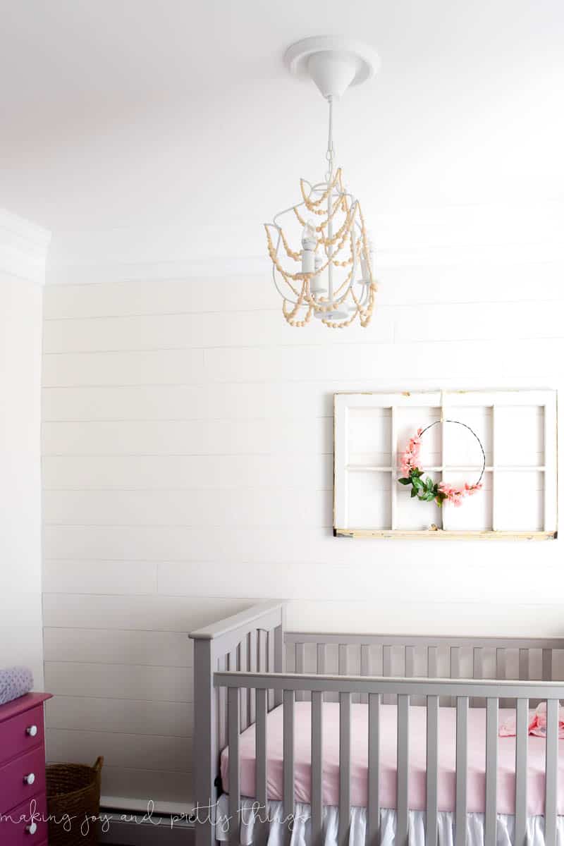 A diy wood bead chandelier hangs over the baby's crib, and farmhouse style wall art hangs on the wall - a vintage window with a white floral hoop.