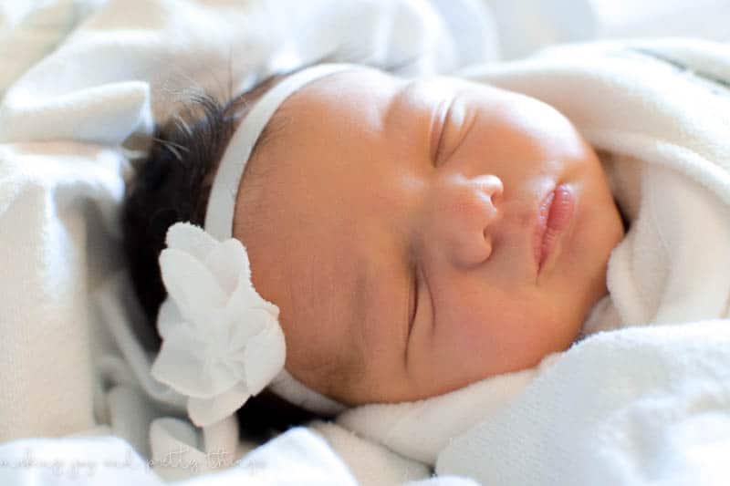 A sleeping newborn baby girl lays wrapped in a swaddle with a white flower headband on her head.