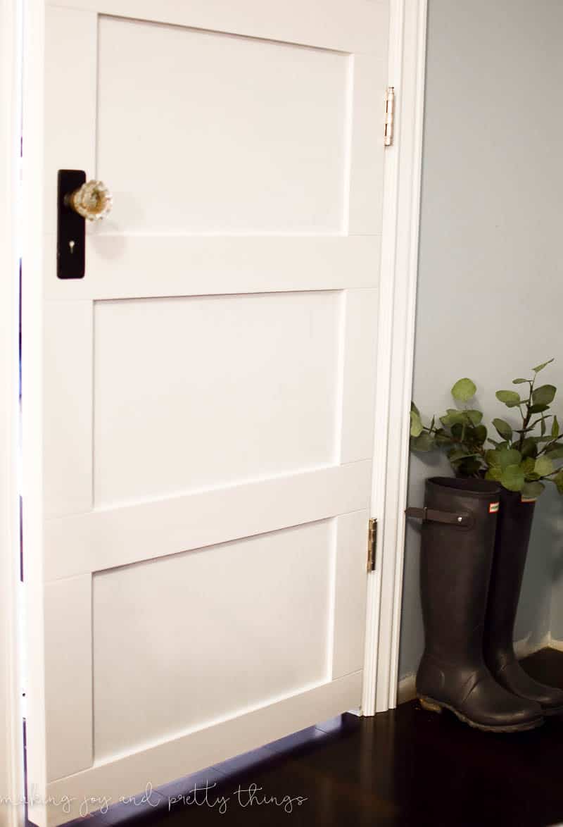Another look at the all-white farmhouse style closet door with antique crystal and brass hardware. A pair of black rainboots filled with greenery sit next to the white closet door.
