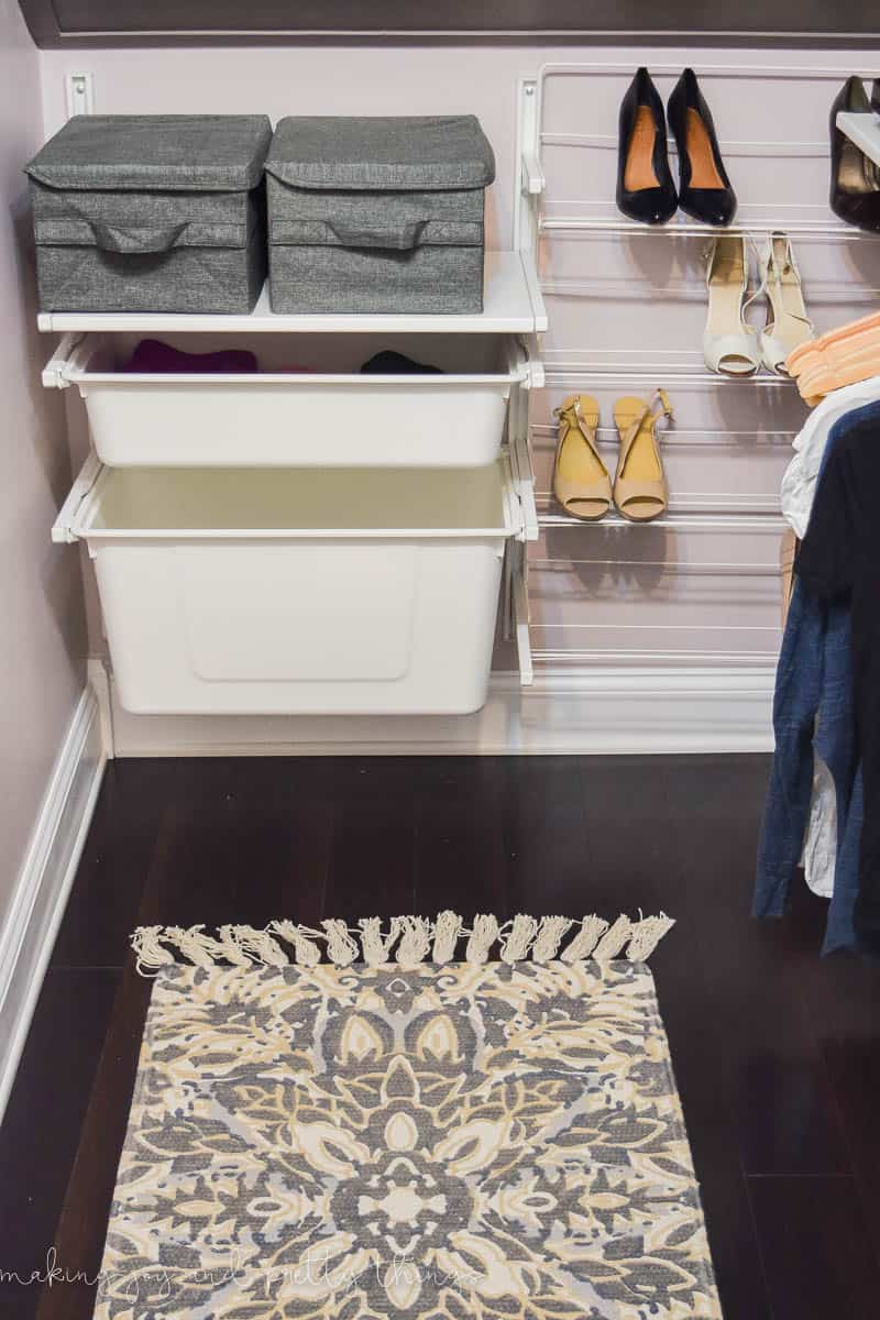 The back wall of our attic walk-in closet is set up with wall-mounted shelving to hold storage bins, baskets, and even a wall-mounted shoe rank. On the floor is a neutural-tone area run with yellow, blue, tan and gray designs with tassels on the end.
