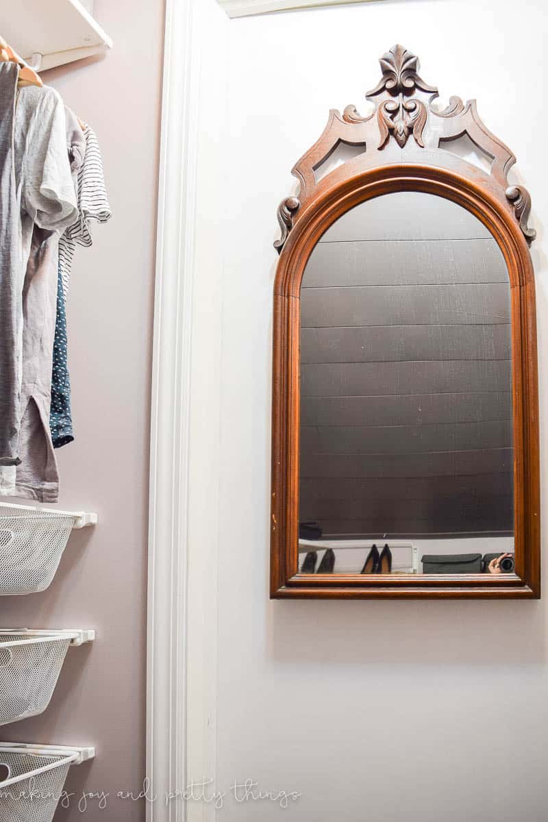 An antique dark wood-framed mirror hangs on the back of a white closet door. The mirror is arch shape with ornate wood crown molding design on top.