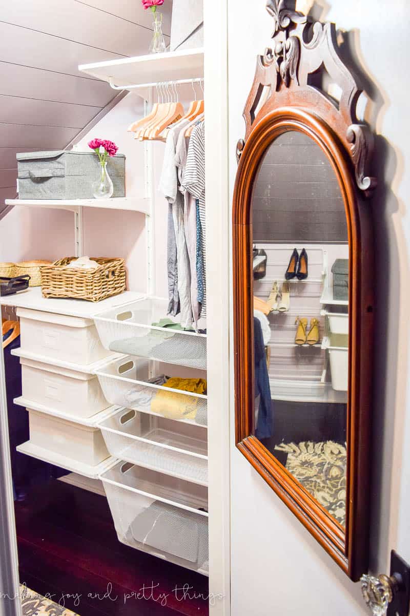 A look inside our updated attic walk-in closet. A dark wood-framed antique mirror hangs on the door, reflecting the back wall in the closet - a shoe rack and storage bins. The inside closet wall has wall-mounted shelving with storage bins and baskets and clothes hanger rods for organized storage.