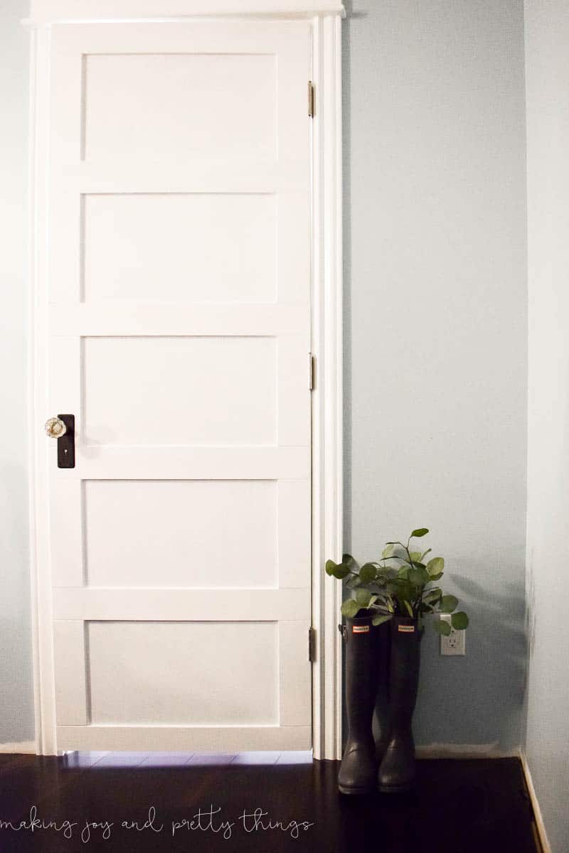 A white farmhouse-style closet door with antique crystal doorknob hardware. A pair of black rubber rainboots filled with faux greenery sit on the floor next to the closet door.