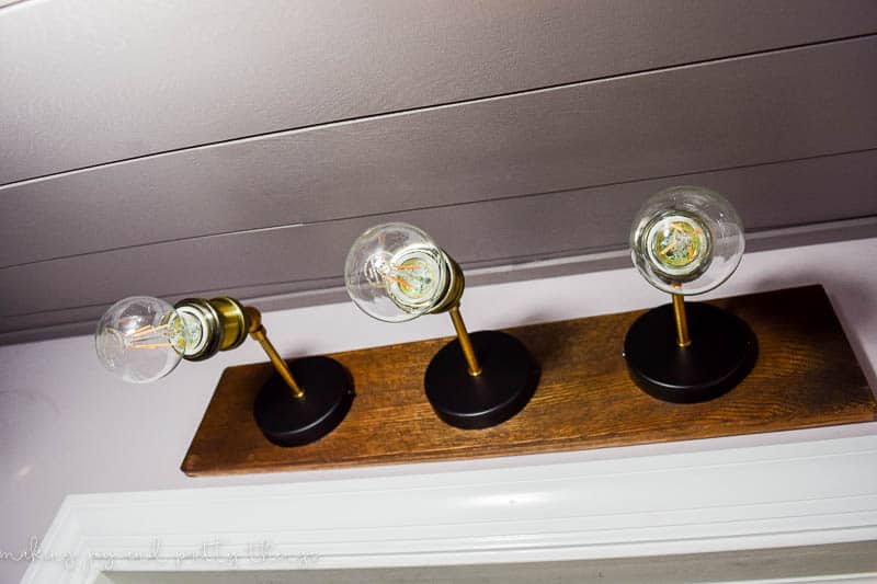 An industrial farmhouse style light fixture attached to the wall above the door in the walk-in closet. The light fixture is a wooden slab with 3 industrial style sconces and clear Edison lightbulbs.