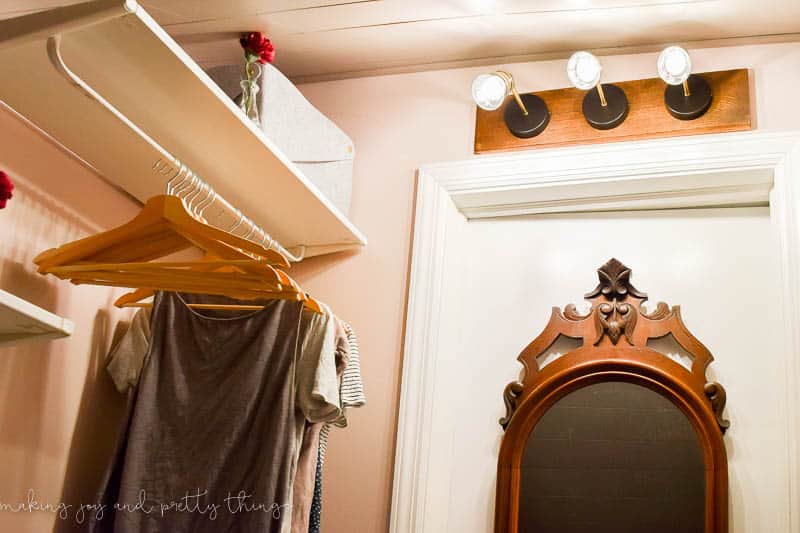 An image shows a lower angle look up at the industrial farmhouse style light fixture in the small walk-in closet. The lights are illuminated, casting light on the wall-mounted shelving and clothing hangers in the closet.