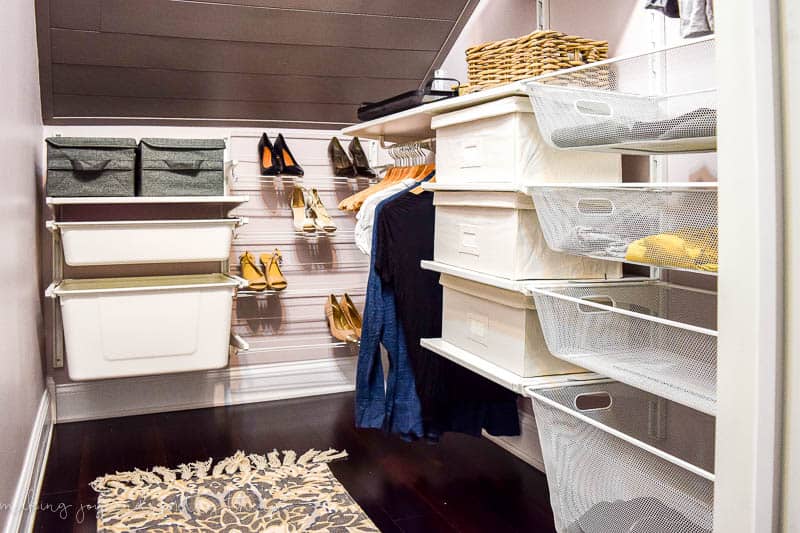 A complete look at our attic walk-in closet storage system. Wall-mounted shelving holds storage bins and baskets for organized clothes storage. Clothing racks with hangers keep clothes organized, and a wall-mounted shoe rack holds women's shoes.