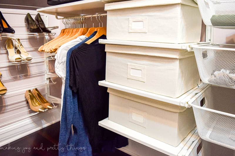 The IKEA Algot closet storage system has wall-mounted shelving to hold storage bins and baskets. Three shelves hold cream colored fabric baskets and white wire baskets for clothes storage. A hanging rod holds wooden hangers and clothes. Against the back wall is a 4-tier shoe rack holding women's shoes.