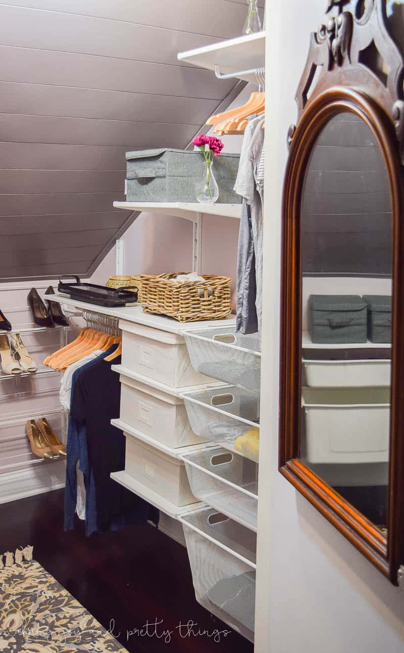 The inside of an attic walk-in close with sloped walls. A wood-framed mirror hangs on the closet door. Wall-mounted shelves hold storage boxes and baskets, as well as two closet rods for hanging clothes. Against the back wall is a4-tiered shoe rack holding pairs of women's shoes.