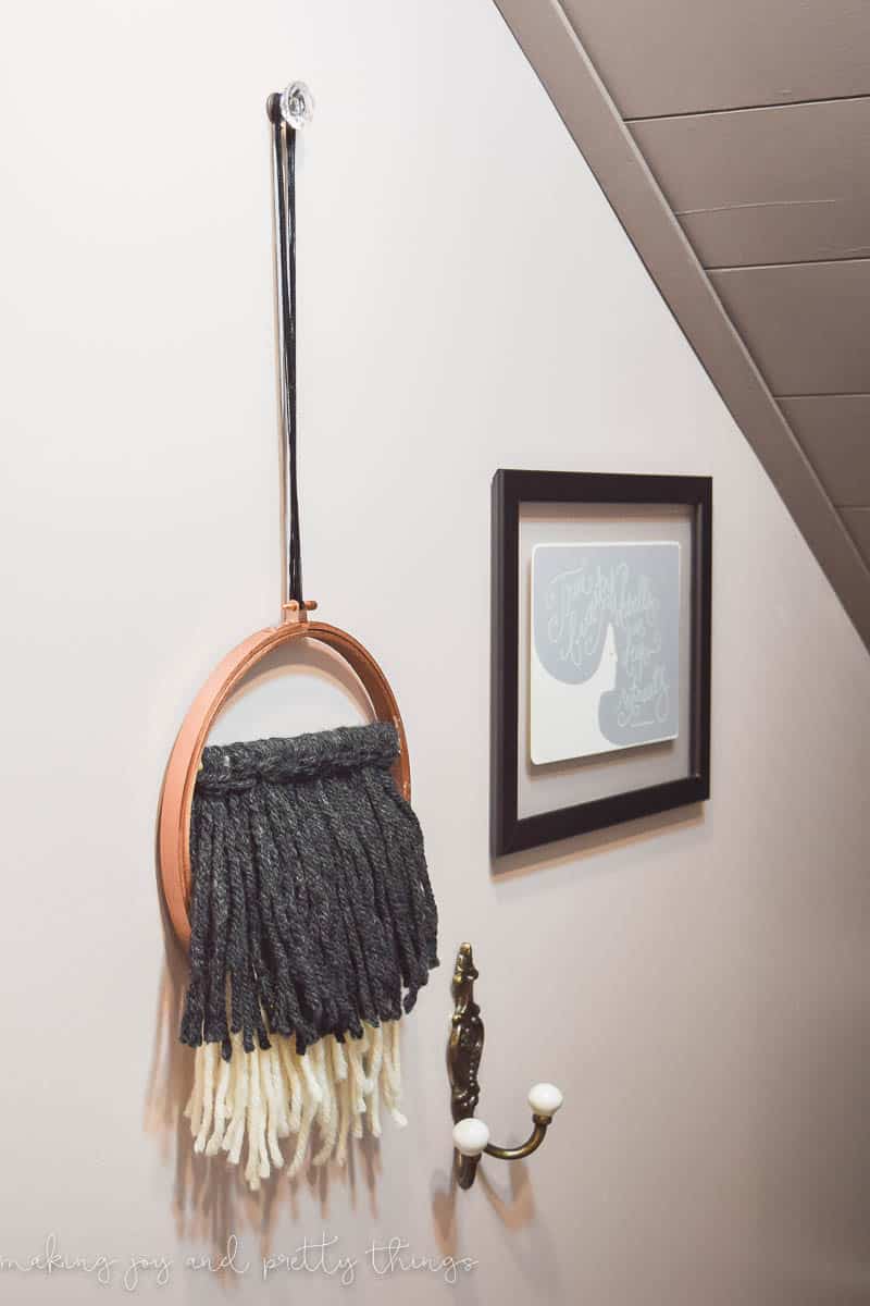 A white wall in the walk-in closet holds a floating black picture frame, an antique wall-mounted, 2-prong coat hanger, and an embroidery hoop wall hanging with tasseled yarn.