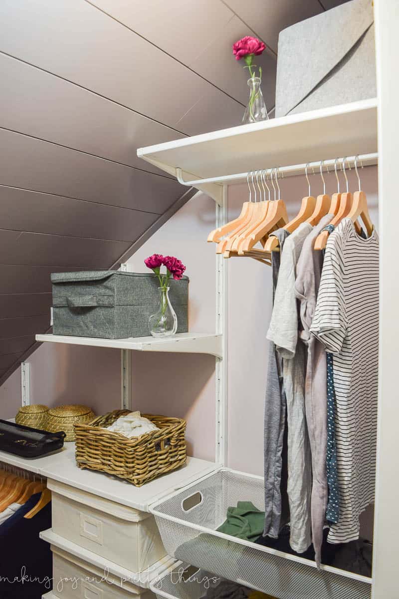 The IKEA Algot closet system installed in our attic walk-in closet. This closet organization system includes wall-mounted shelving that holds storage bins and wire baskets for organization, as well as clothing racks. Clothes hang from wooden hangers, and the shelves hold various storage bins, baskets, and farmhouse-style decor.