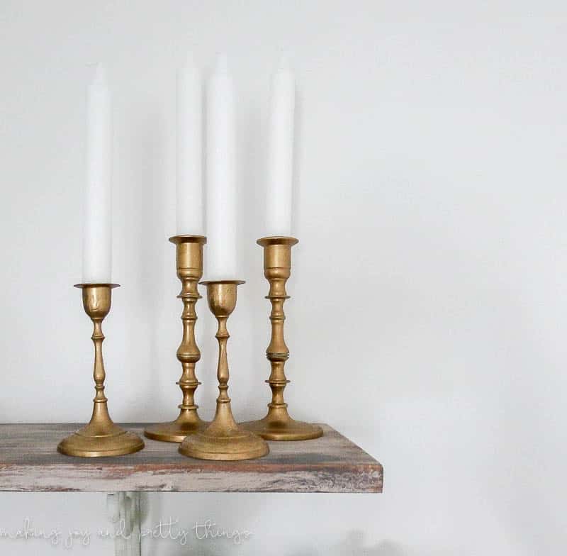 A set of four golf candleholders with while stick candles sit on a wood shelf, a potted ivy plant on the shelf below.