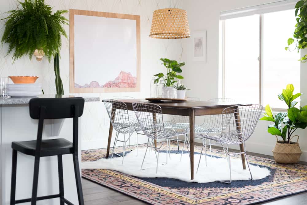 A modern dining room with a wood table and four metal chairs, a plant on the side, then a patterned carpet with a sheepskin rug over the top is on the floor