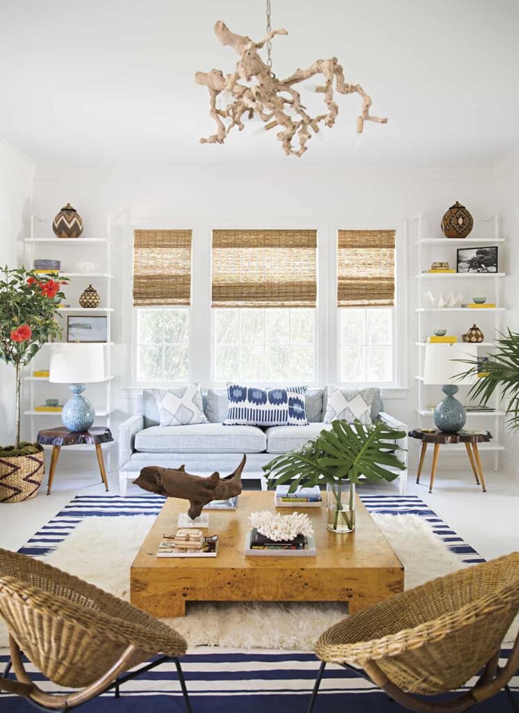 A bright, natural-light filled living room is decorated with coastal style. The centerpiece of the room is a large natural wood table which sits over two large layered area rugs. The room is filled with bright, colorful decor - plants, wicker chairs, sculptures, bookshelves, and more.