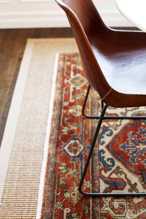 A leather dining room chair sits on layered rugs in a dining room area. The bottom rug is a larger tan woven rug. Layered on top is an ornate oriental style rug with deep reds, blues, and orange colors.