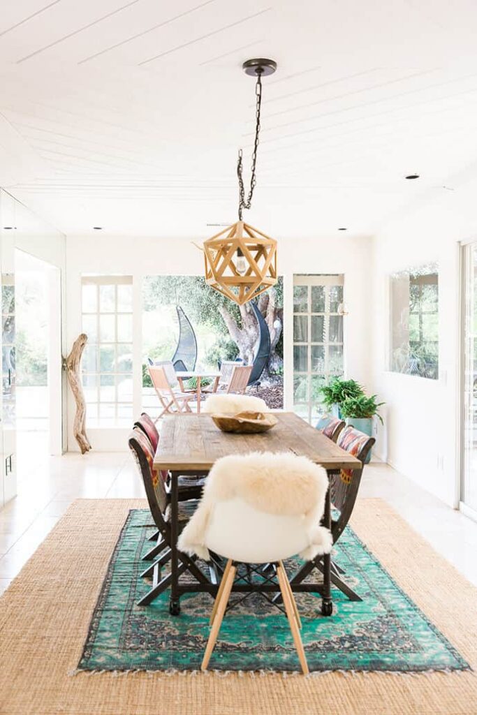 A dining room filled with natural light. A long wooden dining table sits in the middle of the room with six chairs around the table. Two layered rugs sit under the dining room table and chairs.