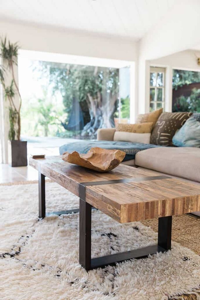 A living room table made from blocks of wood and industrial metal bar legs sits on top of two layered rugs - one shaggy white rug atop a rattan-style woven rug. In the background, a tan couch with throw pillows and floor-to-ceiling windows show a zen outdoors space.