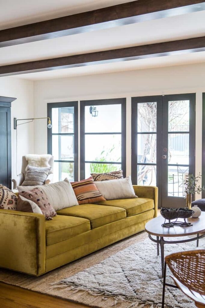 A bright and sunny living room features a large gold velvet couch filled with throw pillows. The couch sits on top of two layered area rugs. An oval coffee table is decorated with contemporary style decor.
