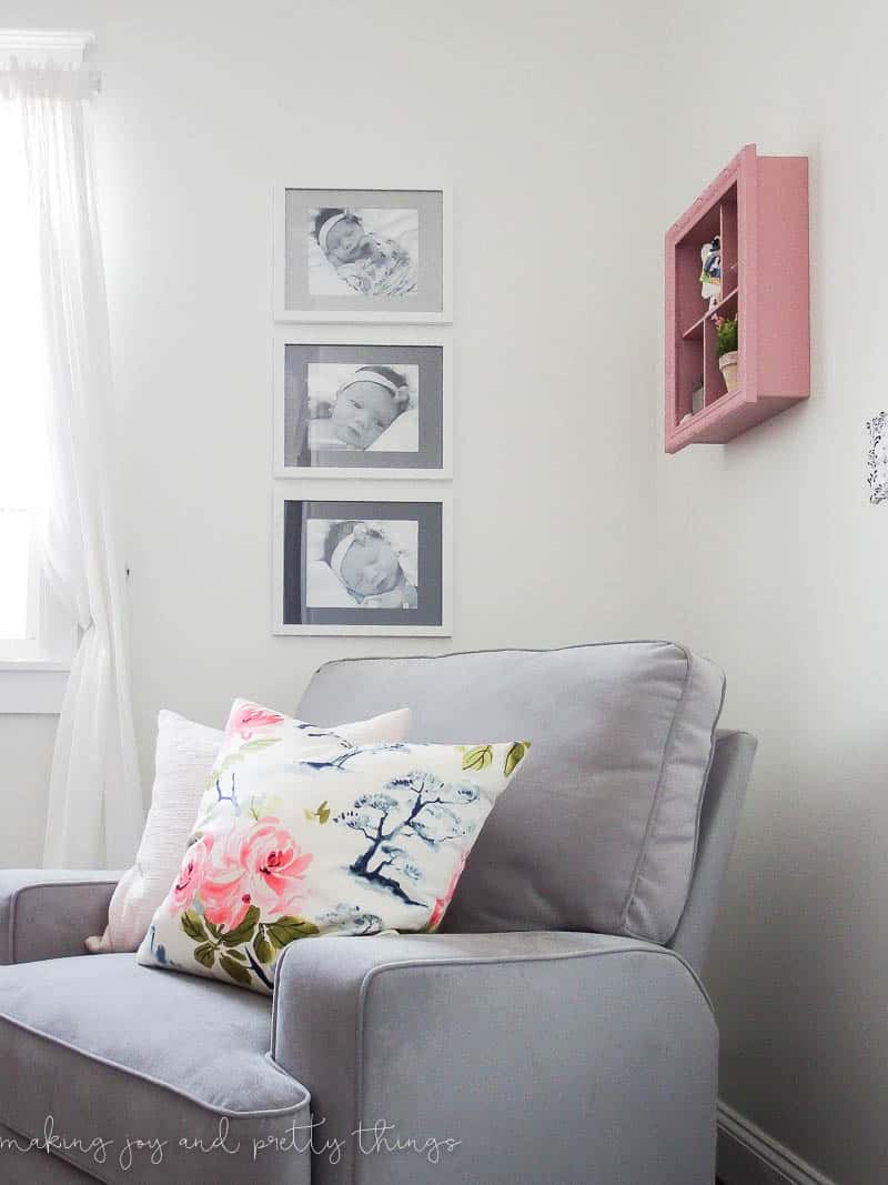 Three framed black and white photos of a newborn baby hang on a white wall, next to a pick shadow box and behind a gray chair with white pillows. The frames have white frames and painted mat boards in three shades of grey.