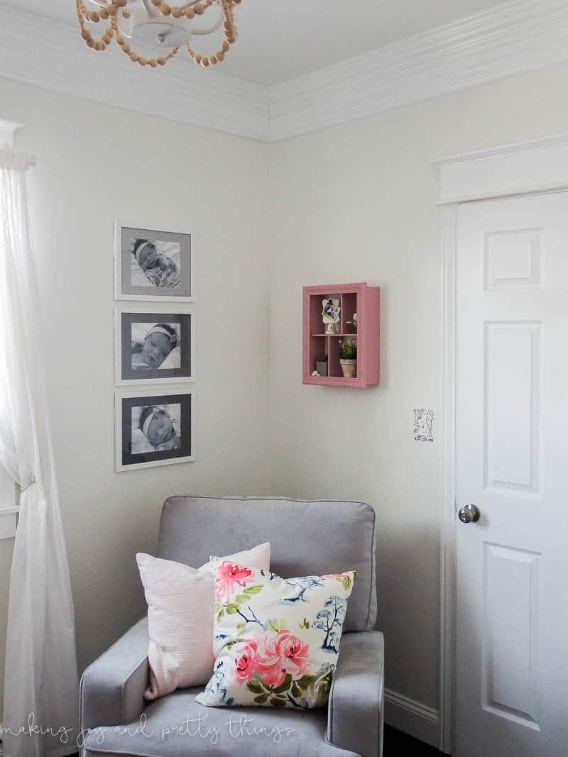 A corner of a baby nursery is decorated with a grey chair with white floral pillows and a pick shadow box on the wall. On the other wall hangs three picture frames in a vertical line. Each frame holds a black and white photo of a baby, with a painted mat board.