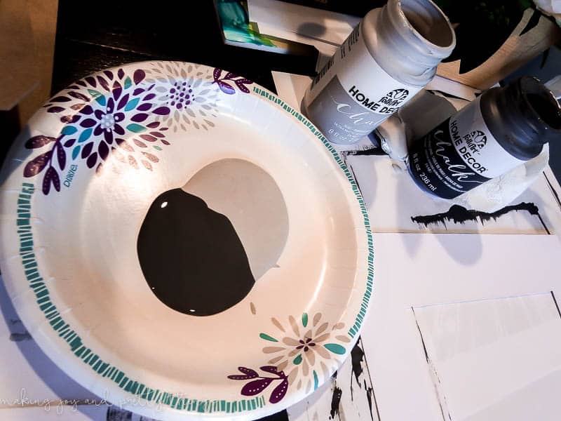 Two pools of chalk paint - black and grey - floor together in a paper bowl with a flower design. This mixed paint color will be used for painting mat boards, giving them an  ombre color effect.