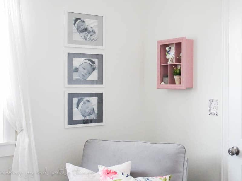 Three framed baby photos hang on a wall next to a window and a pink shadow box. The picture frames hold back and white baby photos, and the mat boards are painted different shades of grey to create an ombre effect.