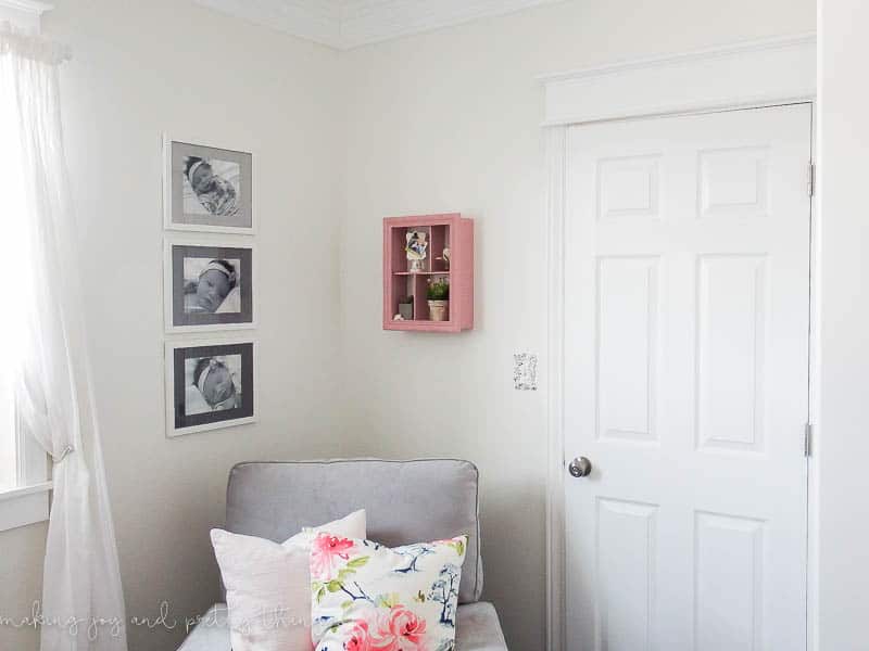 A corner of a baby's nursery. There is a white door, a pick shadow box hanging on a wall, a grey chair with white and floral-patterned pillows, and three framed baby photos with painted mat boards in shades of grey.