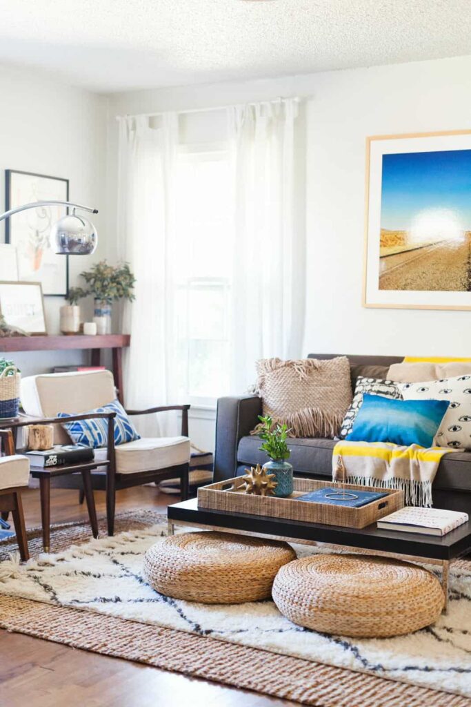 A welcoming and modern living room. A charcoal grey couch is decorated with colorful throw pillows. Two white linen arm chairs sit side-by-side with a small side table between them. A low-to the ground coffee table holds a large serving tray filled with decor. Two plush seat cushions sit under the coffee table, atop two layered area rugs.