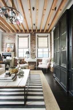 A very modern-style living room with exposed wood beam sealings, a dark accent wall, white linens arm chairs, and a decorated wood table with metal frame legs. There is a large woven area rug on the floor, with a smaller blue and white striped area rug layered on top.