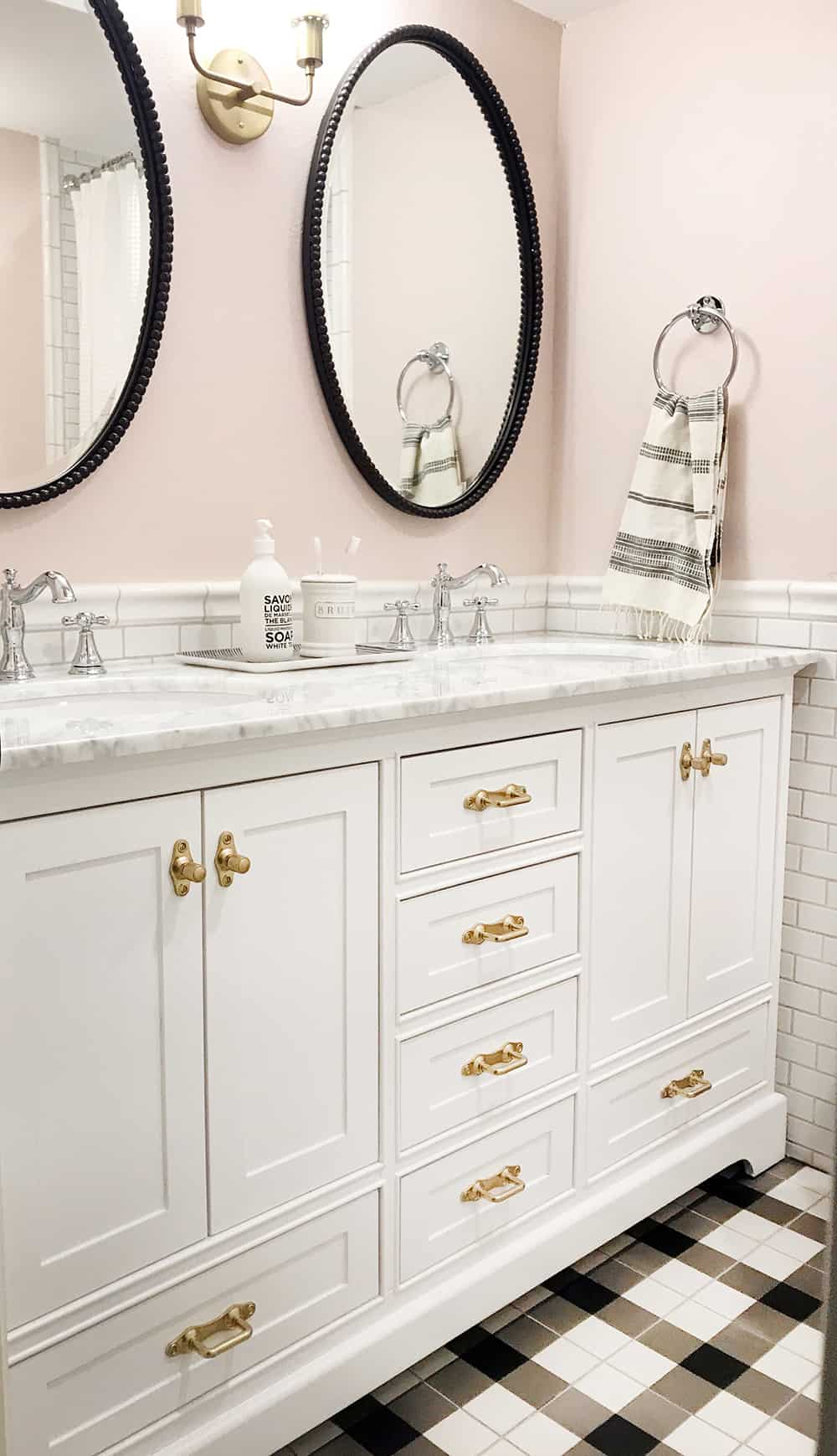 A modern vintage bathroom with white dual sink vanity, white cabinets with gold handles, and white marble countertops. The walls are half white subway tiles, and half painted a light blush pink. Two metal framed oval mirrors hang above each sink.